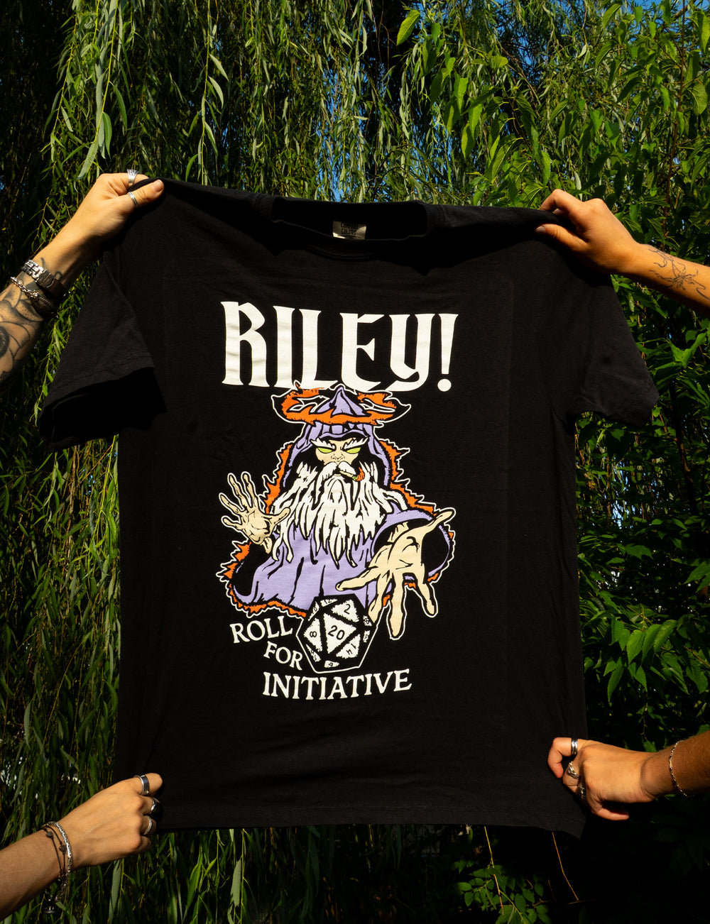 A black printed t-shirt from Riley!, held by four hands, features a wizard, a 20-sided die, and the text "RILEY! ROLL FOR INITIATIVE," set against a backdrop of green plants and sunlight. Crafted on Comfort Colors fabric, this tee is perfect for tabletop gaming fans.