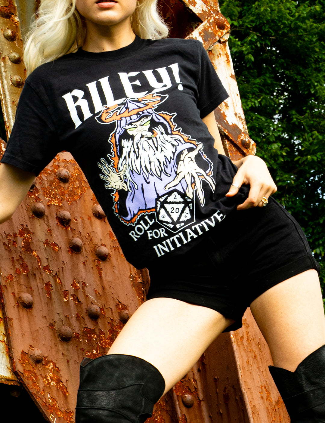 A person in black shorts and boots wears a Riley! Comfort Colors printed t-shirt featuring a wizard, a 20-sided die, and the text “ROLL FOR INITIATIVE,” standing outdoors by a rusty metal structure.