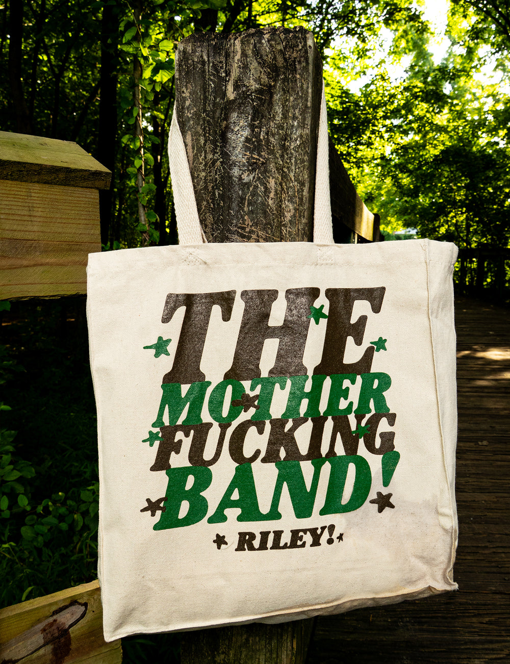 A canvas tote bag hangs on a wooden post, displaying the text THE MOTHER FUCKING BAND! Riley! in bold black and green letters. The background features a sunlit forest and wooden path, with the brand name Riley! prominently shown.