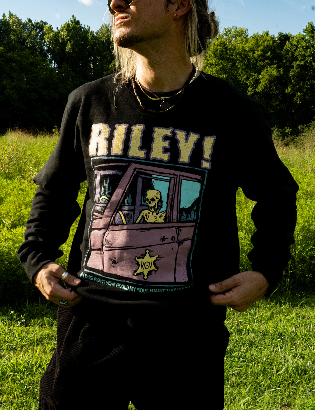 A person stands outdoors in a black Riley! sweatshirt featuring an illustration of a skeleton driving a car, with a green field, trees, and blue sky in the background.