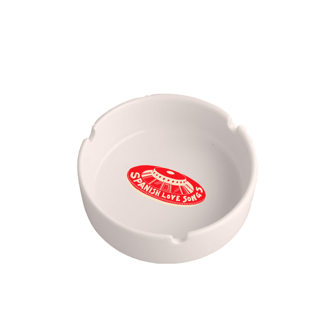 A white ceramic ashtray with three grooves, featuring the Spanish Love Songs logo in red and white and an illustrated record at the center.