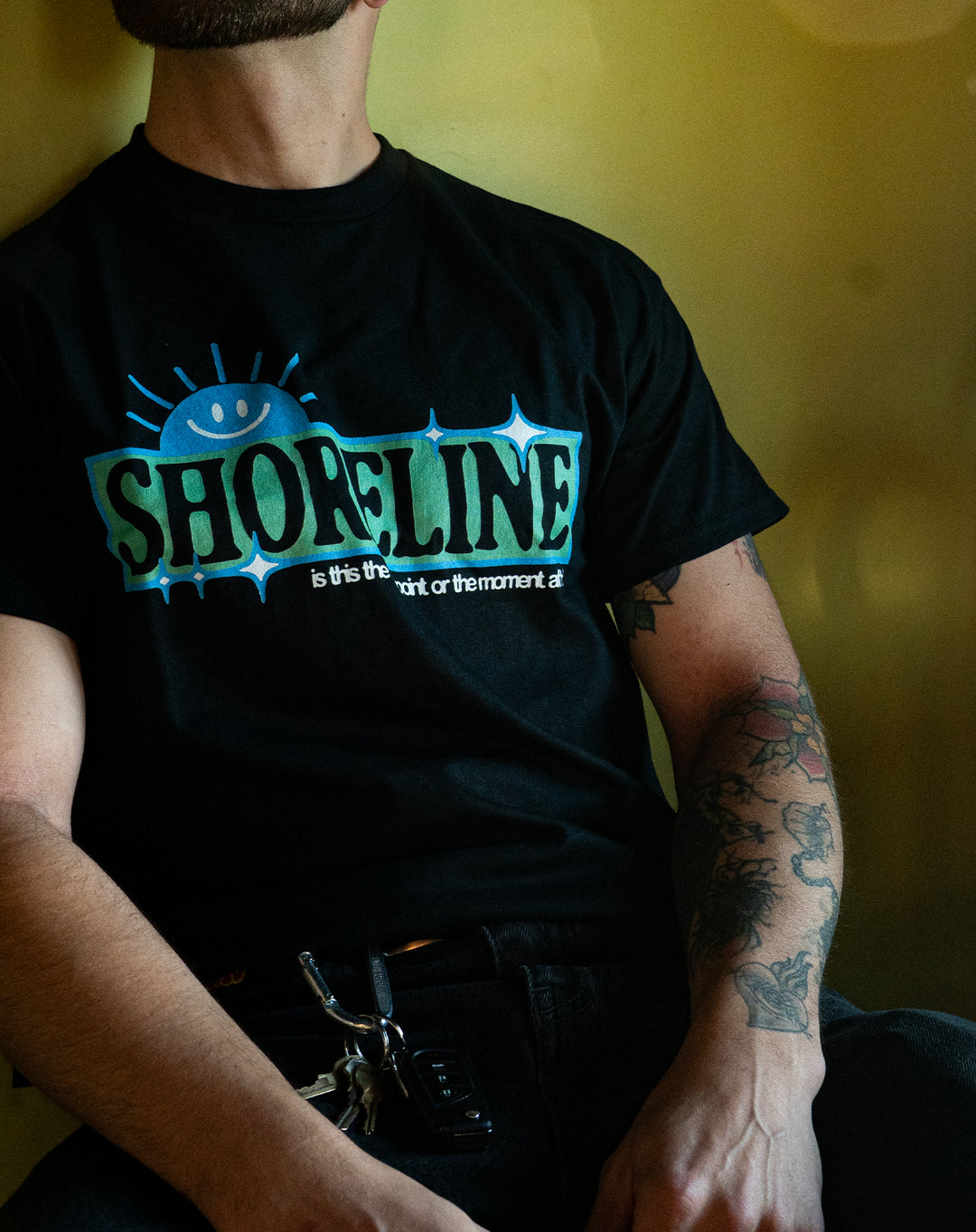 A person with a partial beard and tattooed arm sits against a yellow background, wearing a black Shoreline shirt featuring "SHORELINE" with a blue smiling sun graphic and small text below, plus keys clipped to their belt loop.