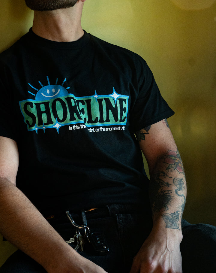 A person with a partial beard and tattooed arm sits against a yellow background, wearing a black Shoreline shirt featuring "SHORELINE" with a blue smiling sun graphic and small text below, plus keys clipped to their belt loop.