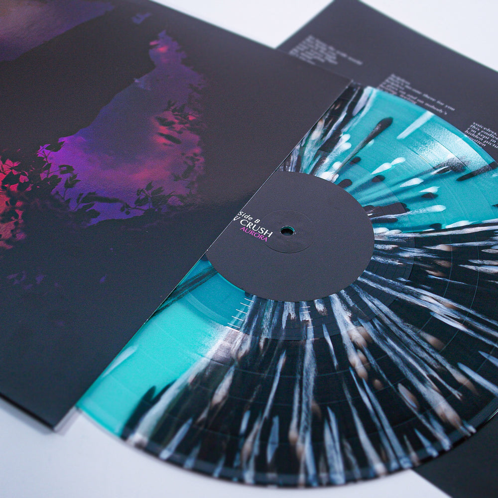 A vibrant vinyl from Slow Crush with a black center label and teal, white, and black splatter slides partway out of its moody album cover, while another sleeve featuring white text rests in the background.