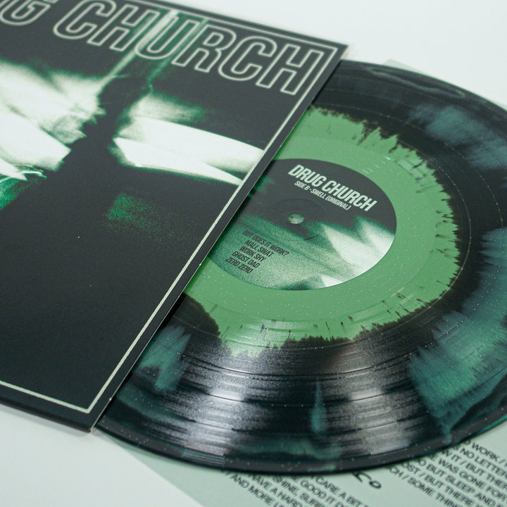 A marbled Mint/Black/Olive vinyl record by Drug Church is partially pulled from its album cover, showing bold white "DRUG CHURCH" text. The label and inner sleeve are visible. Release date: June 5th, 2026.