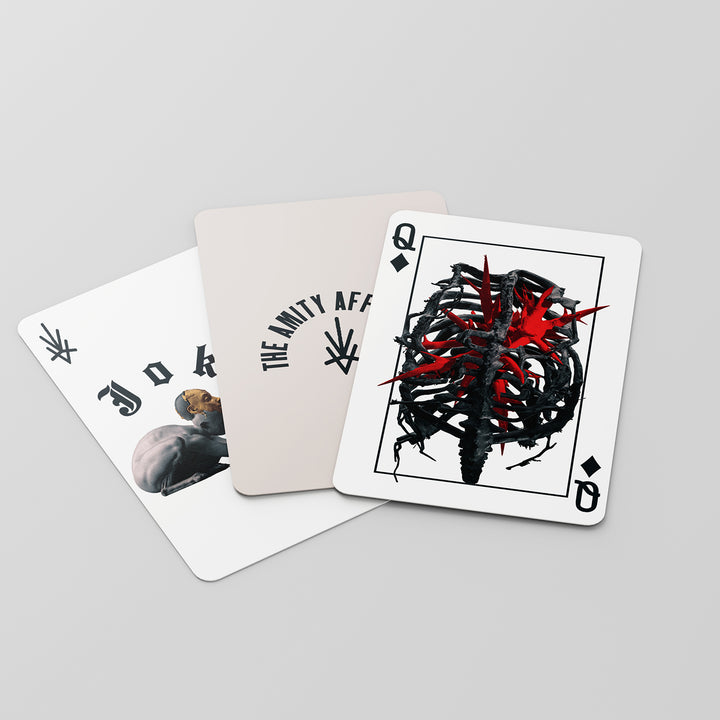 Three playing cards on a light surface: a Joker card with a resting buffalo, a The Amity Affliction branded card, and a Queen of Spades card featuring a thorny heart pierced by red spikes.