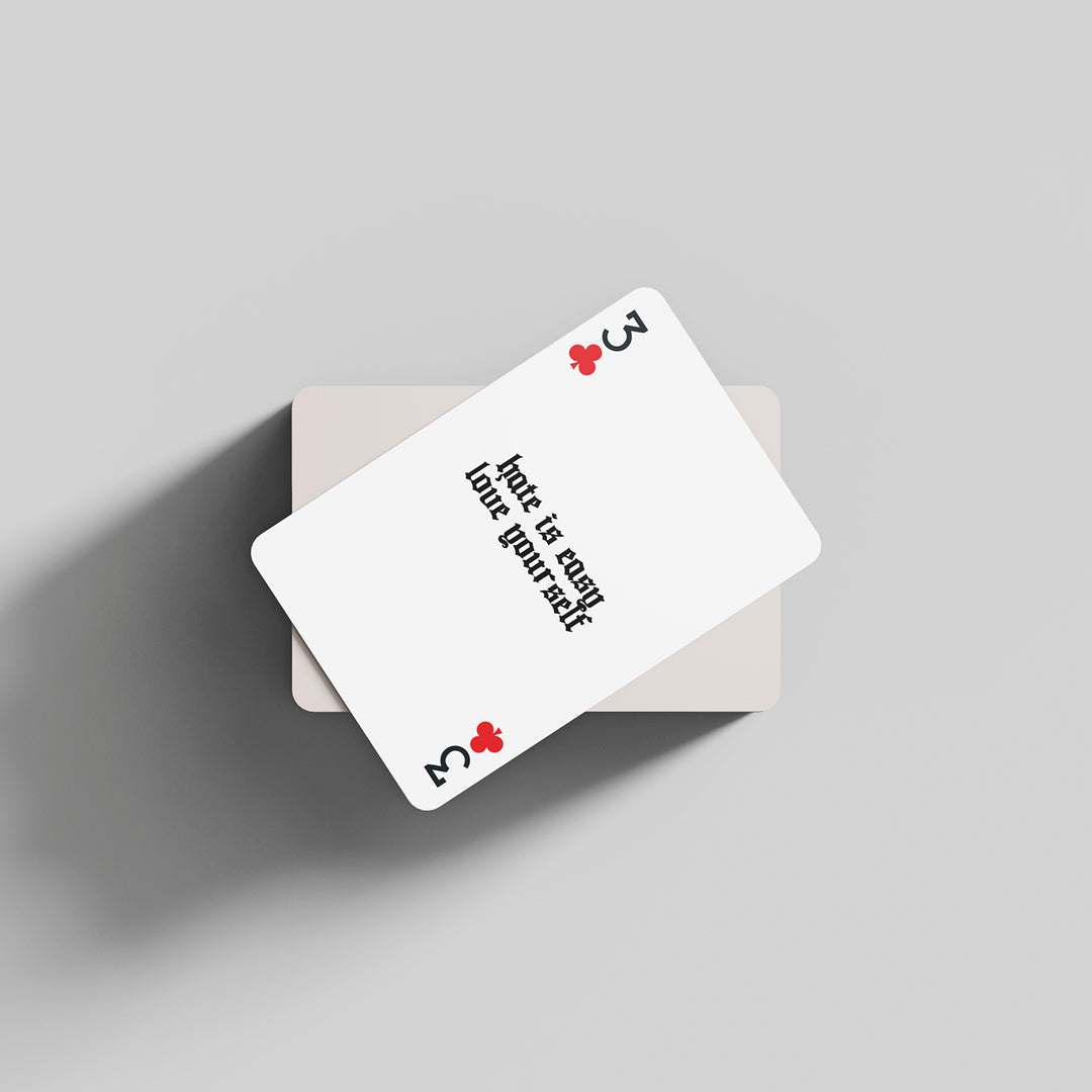 A three of clubs playing card by The Amity Affliction with "Hate is easy, love yourself" in black text, resting at an angle on a face-down deck against a light gray background.