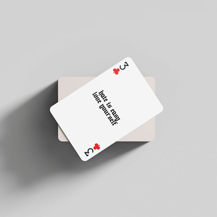A three of clubs playing card by The Amity Affliction with "Hate is easy, love yourself" in black text, resting at an angle on a face-down deck against a light gray background.