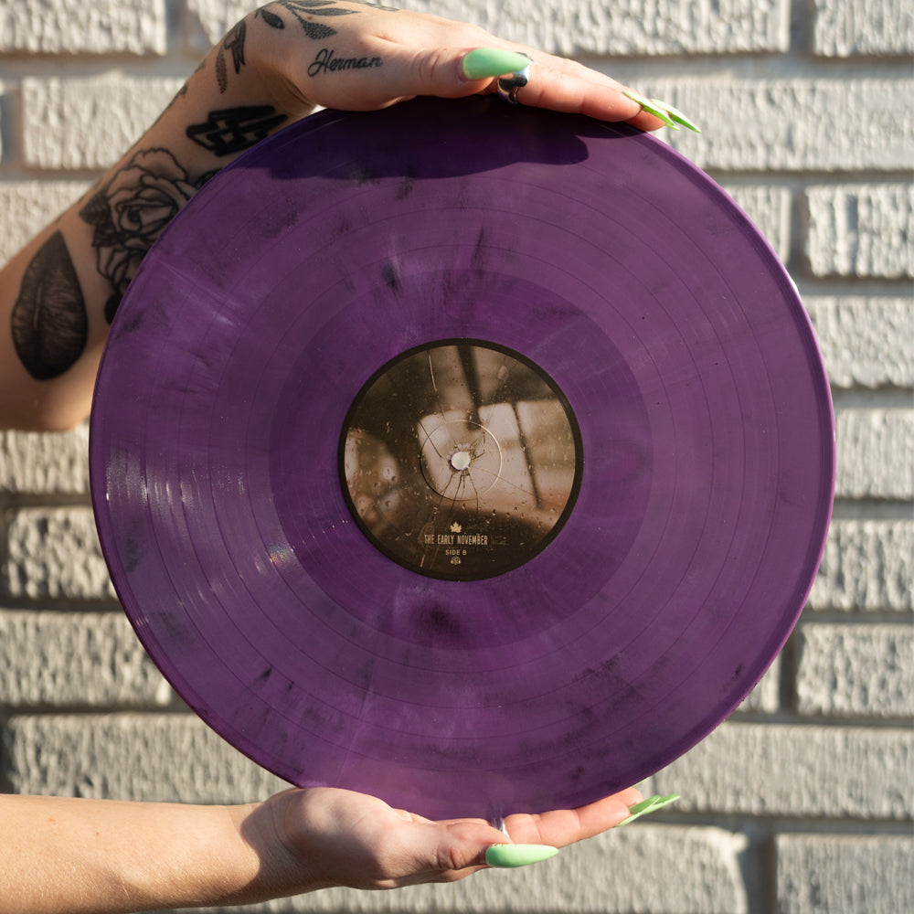 A person with tattoos and long green nails holds The Early November Eco Mix Vinyl LP before a white brick wall. Sunlight highlights the translucent purple record—an eco-friendly must-have for collectors and fans of The Early November.