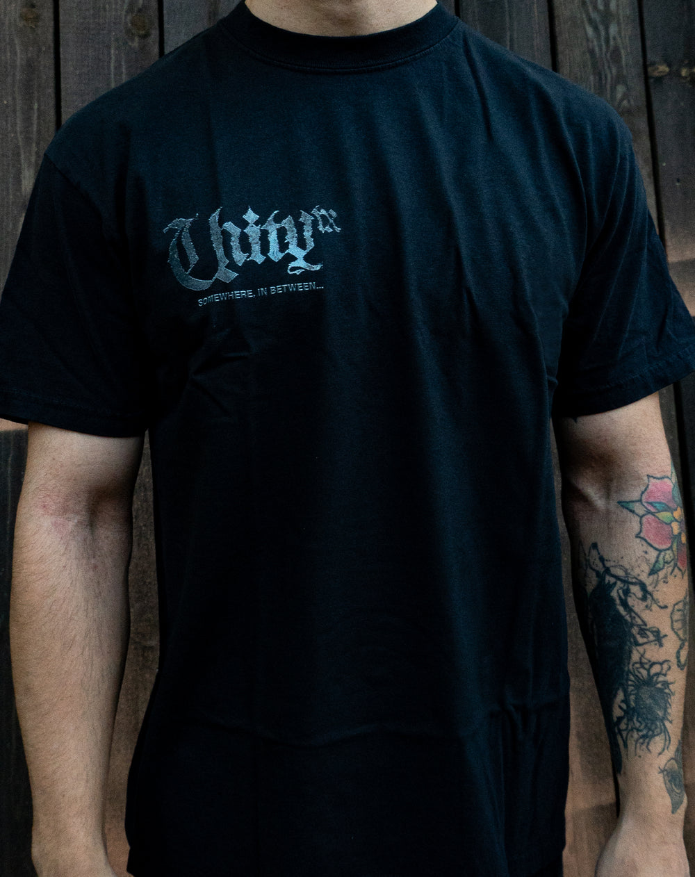 A person wearing a black UNITYTX Comfort Colors tee with "UNIVR" and "SOMETHING...IN BETWEEN..." in white on the chest. Their tattooed right forearm is visible as they stand by a wooden wall.