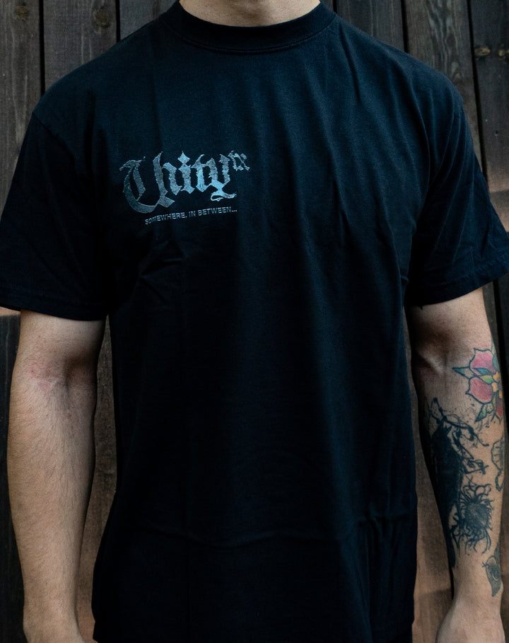 A person wearing a black UNITYTX Comfort Colors tee with "UNIVR" and "SOMETHING...IN BETWEEN..." in white on the chest. Their tattooed right forearm is visible as they stand by a wooden wall.