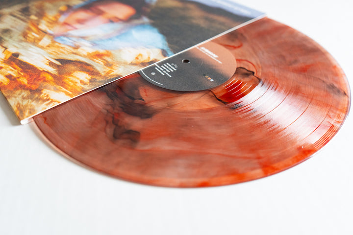 A close-up of a Shoreline Amber Deluxe Marble Vinyl LP, partially out of its cover displaying blurred warm-toned artwork. This vinyl LP presale is elegantly presented on a white surface.