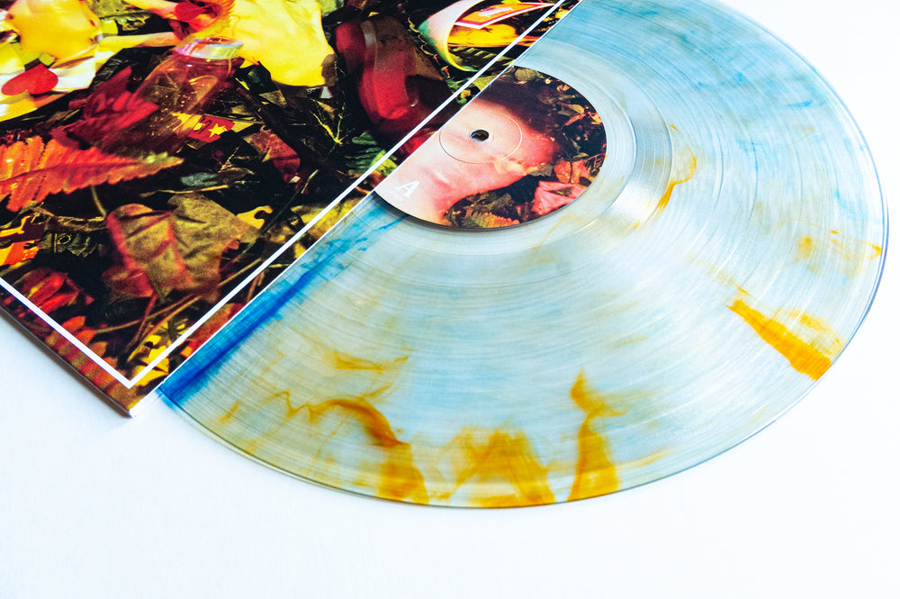A transparent Drug Church Marble Vinyl with blue and yellow streaks is partially pulled from the Hit Your Head LP cover featuring vibrant objects, resting on a white surface for the exciting Album Release 2026.