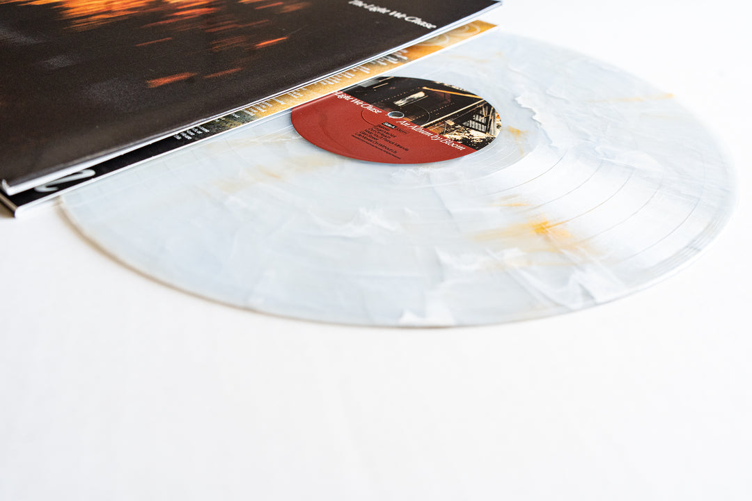 A marbled white Bloom Golden Pearl Vinyl LP with faint gold streaks is partially slipped from its cover, which displays a blurred cityscape, all set on a white surface—ideal for any album pre-order.