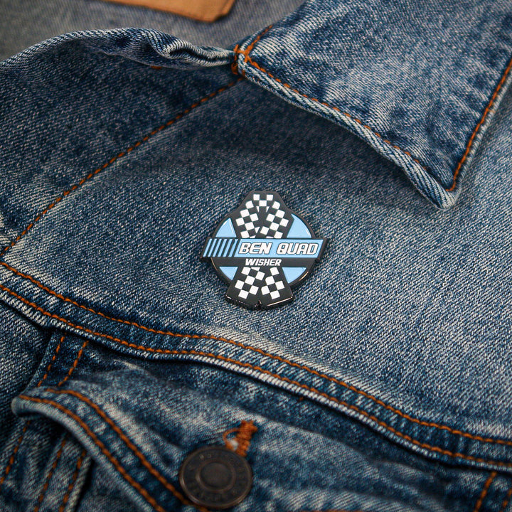 A diamond-shaped enamel pin with a black-and-white checkered pattern and blue text reading BEN QUAD WISHER pinned to a blue denim jacket.