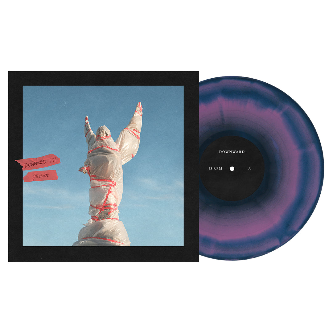 A DOWNWARD Deluxe Vinyl LP in striking purple and black marbled design sits beside the album cover, which shows a statue-like figure wrapped in plastic and red tape with raised arms against a clear blue sky.