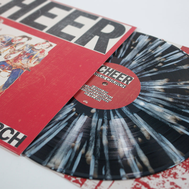 A limited edition black and white splatter vinyl by Drug Church, partially pulled from a red cover with bold CHEER text. Both the record and cover showcase artistic designs and stylized illustrations inspired by Black Ice.