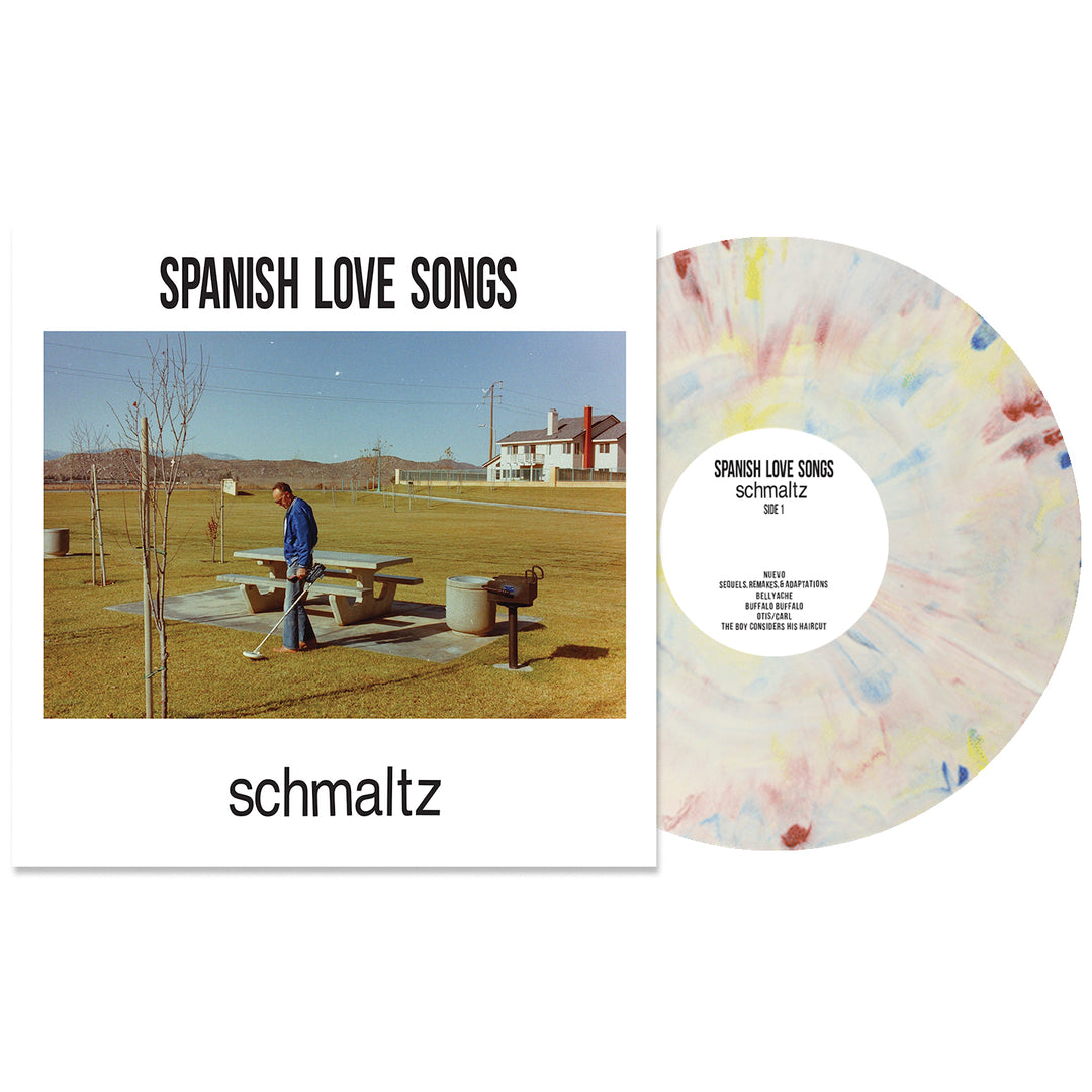 The Schmaltz album cover by Spanish Love Songs shows a person at a picnic table in a grassy park with the Schmaltz Crayon Marble Vinyl LP, its surface featuring colorful pastel splatters, placed beside them.