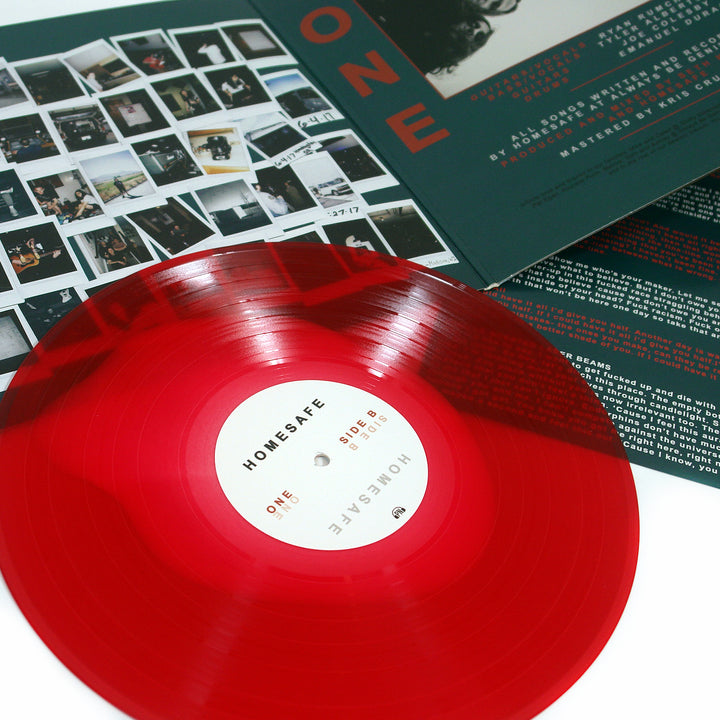 A translucent blood-red Homesafe vinyl record rests on a cream-white surface, accompanied by its album cover and an insert displaying many small photos and lyrics.