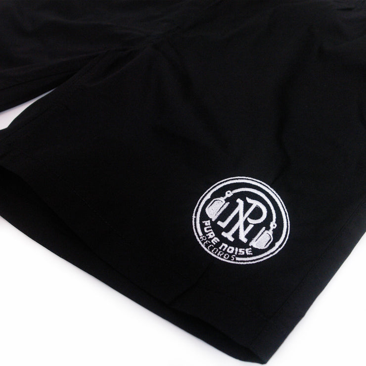 close up image of a pair of black beach shorts on a white background. there is a small embroidered circle on the bottom right showcasing pure noise records logo on the letters P N with headphones around and arched up pure noise records below.