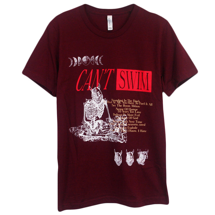 image of a maroon tee shirt on a white background. tee has full body print. in white at the top left are the phases of the moon. below in orange says can't swim, below in the center of the tee on the right in white is a demon skeleton sitting crossed legged and the tracklist to the album Change of Plans is on the right in yellow. On the bottom right are three demon skull heads in white