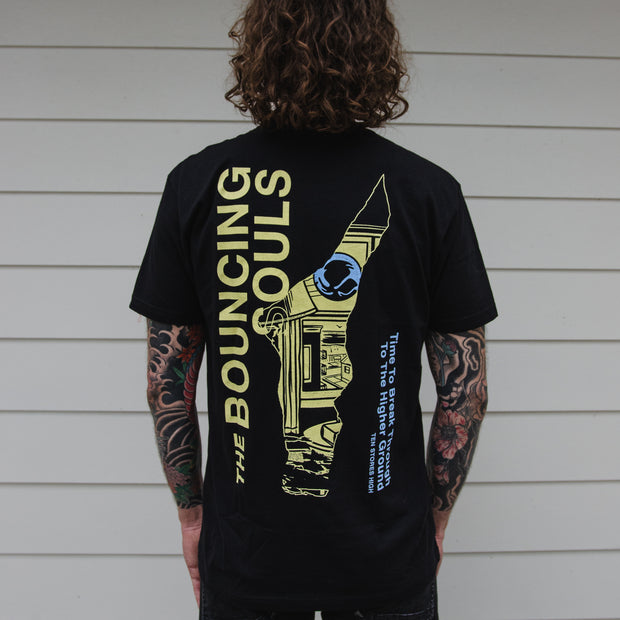 image of a man with tattooed arms wearing a black tee shirt. the back has "The Bouncing Souls" text in yellow, "Time to break through the higher ground, Ten Stories High" text in blue. And a odd shaped cut out in the center of the back with the album art peering through