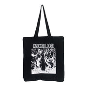 image of the front of a black canvas tote bag on a white background. tote bag  has a full print in white. Knocked loose is written at the top and below that is an image of a persons face with sunglasses on and hands up with white scribbles surrounding it.