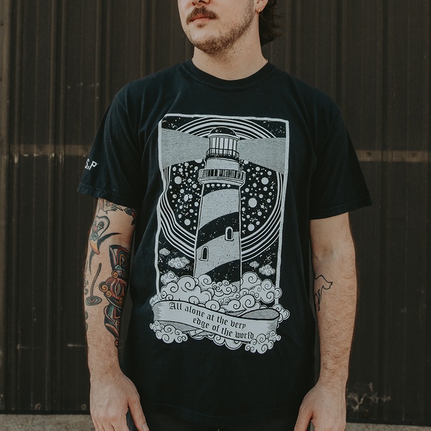 man wearing black tee shirt. black t-shirt with a large lighthouse printed in the center on the tee. clouds at the bottom that have the text All Alone at the very edge of the world. BSP printed on the right sleeve. 