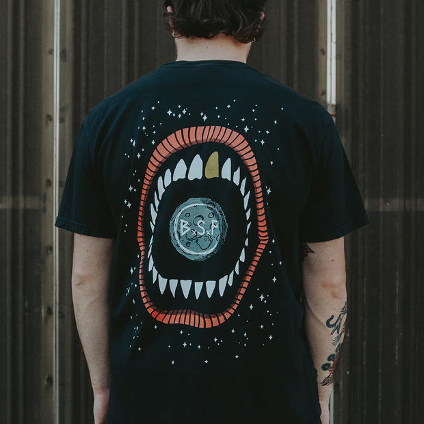 image of the back of a man wearing a black tee shirt. Large mouth with 1 gold tooth and a moon in the center printed on the back with a stary galaxy.