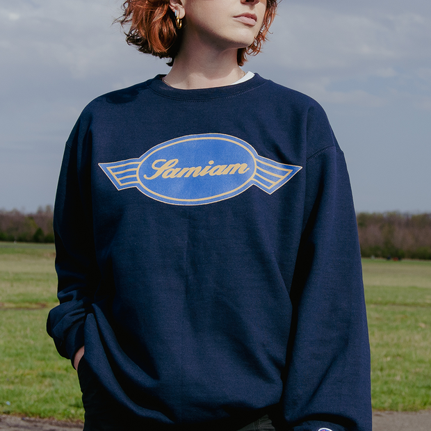 image of a girl outside wearing a navy crewneck sweatshirt.  Crewneck has crest logo printed on the front chest in gold and blue ink. 