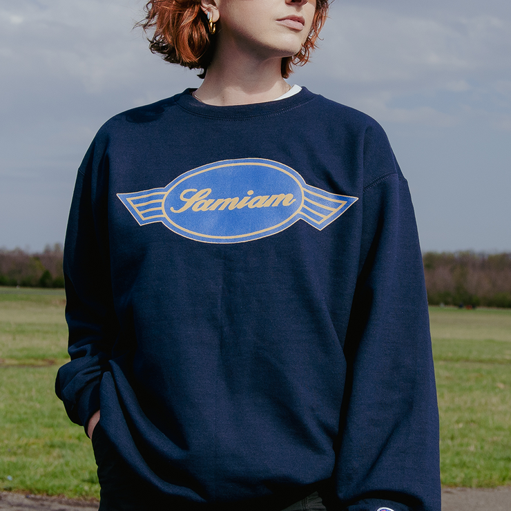 image of a girl outside wearing a navy crewneck sweatshirt.  Crewneck has crest logo printed on the front chest in gold and blue ink. 