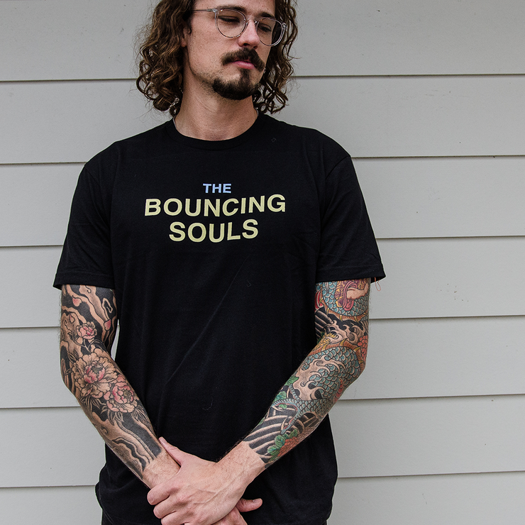 image of a man with tattooed arms wearing a black tee shirt. front has the text "The Bouncing Souls" in the center chest in yellow and blue ink.