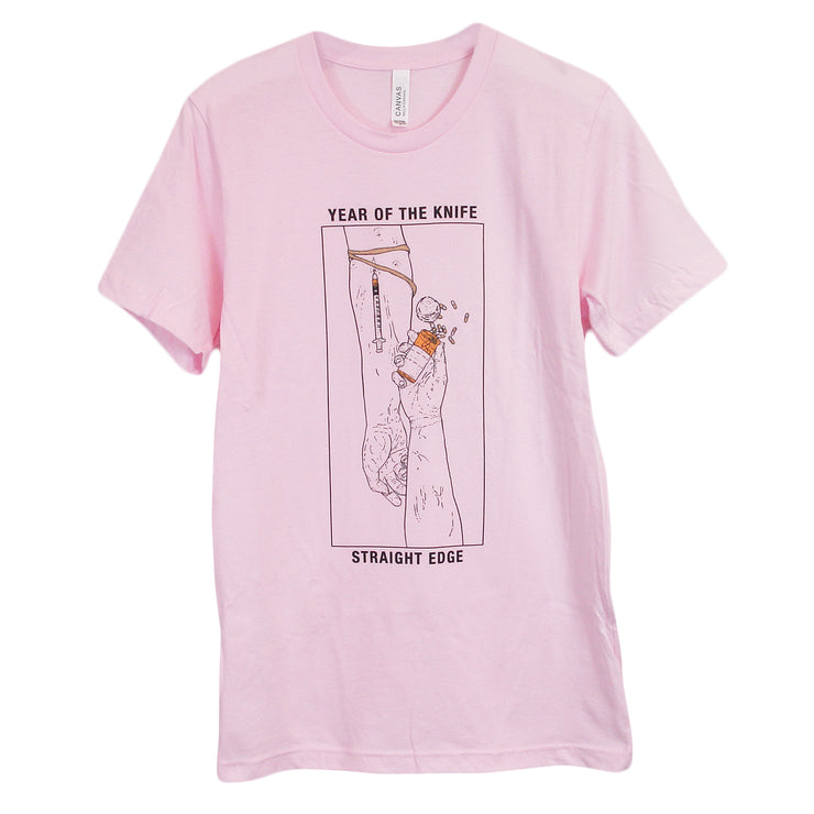 image of a light pink tee shirt on a white background. tee has a full body center print that has a vertical rectangle with two hands. one with a syringe in the arm, the other hand is holding a bottle of pills. on the top of the rectangle says year of the knife and the bottom across says straight edge