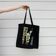 hand holding black tote bag, Bouncing Souls Break Through Black tote bag "The Bouncing Souls" text in yellow, "Time to break through the higher ground, Ten Stories High" text in blue. And a odd shaped cut out in the center of the tote bag with the album art peering through