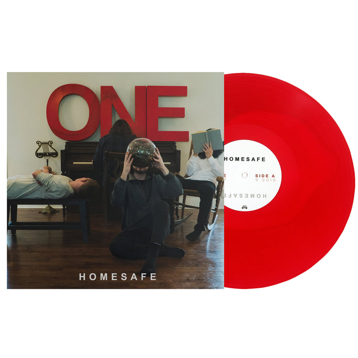 The Homesafe album cover shows three people in a cream room with a piano and bold red ONE lettering. One sitter wears a glass bowl on their head, another reads, and one lies on the piano. A red vinyl record sits nearby.